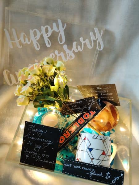 Decorative gift box with flowers, cards, and a mug on a reflective surface with 'Happy Anniversary' text.