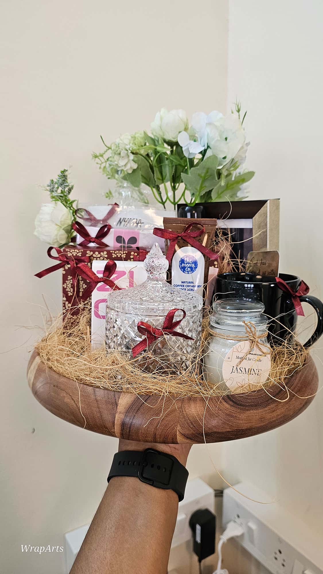 Handheld wooden tray with gift items including a mug, candles, and flowers on a neutral background.