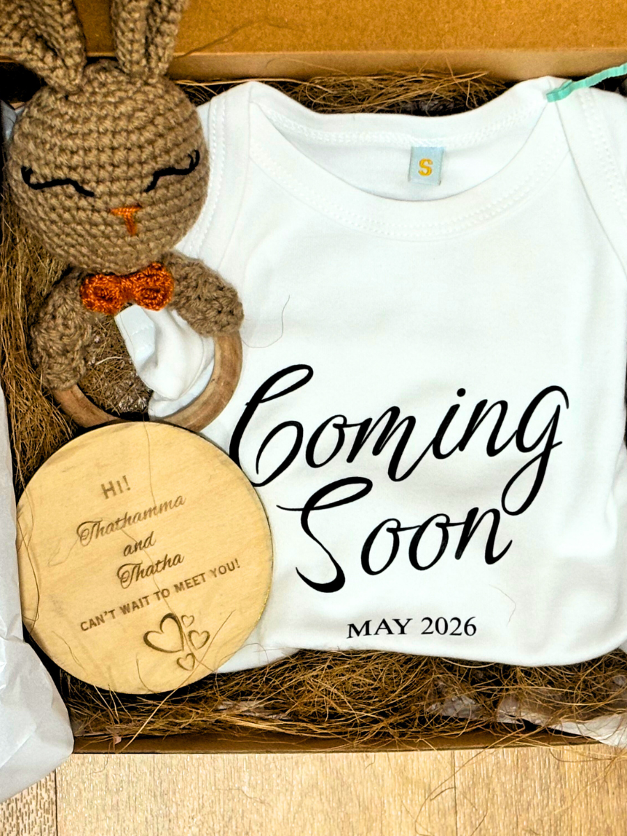 White baby onesie with 'Coming Soon' text, brown crocheted bunny toy, and wooden disc in a box.
