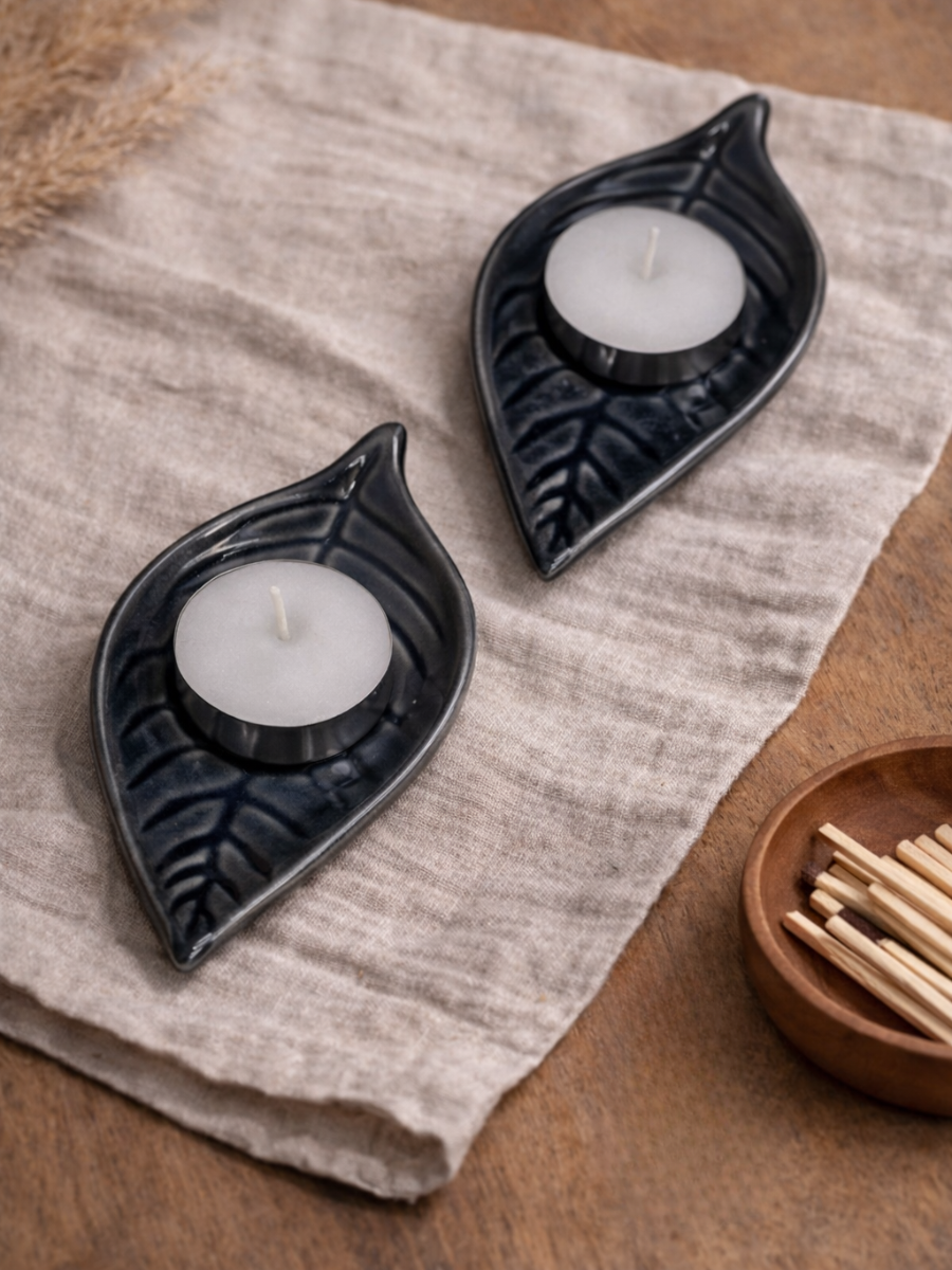 Two black leaf-shaped candle holders with white candles on a textured fabric surface.