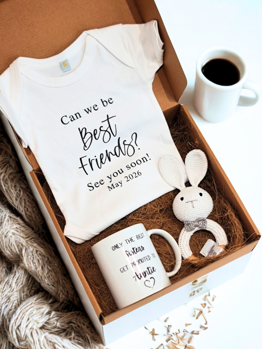 Baby gift set with onesie, mug, and toy in a box on a textured surface.