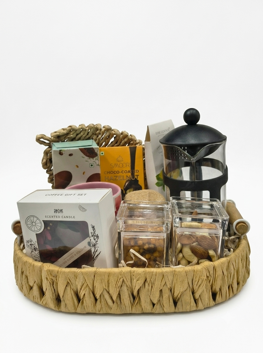 A round woven tray gift set filled with assorted items including a scented candle box, a black French press, glass jars of nuts and dried fruits, and other packaged goods, photographed on a clean white background.