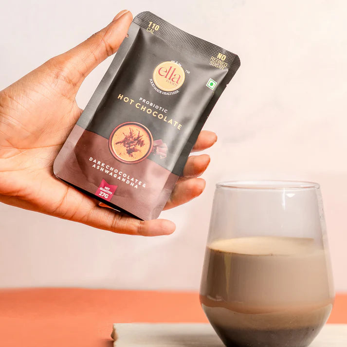 Hand holding a package of Elti Probiotic Hot Chocolate with a glass of hot chocolate in the background.