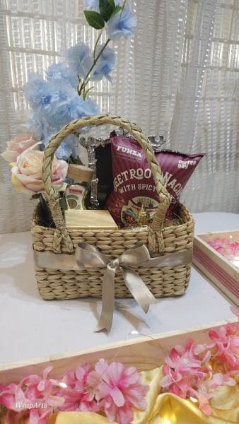 Wicker gift basket with flowers and snacks on a white surface with floral decorations.