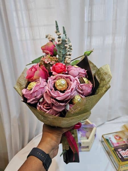Bouquet of pink flowers with chocolate candies held by a person against a white curtain background.